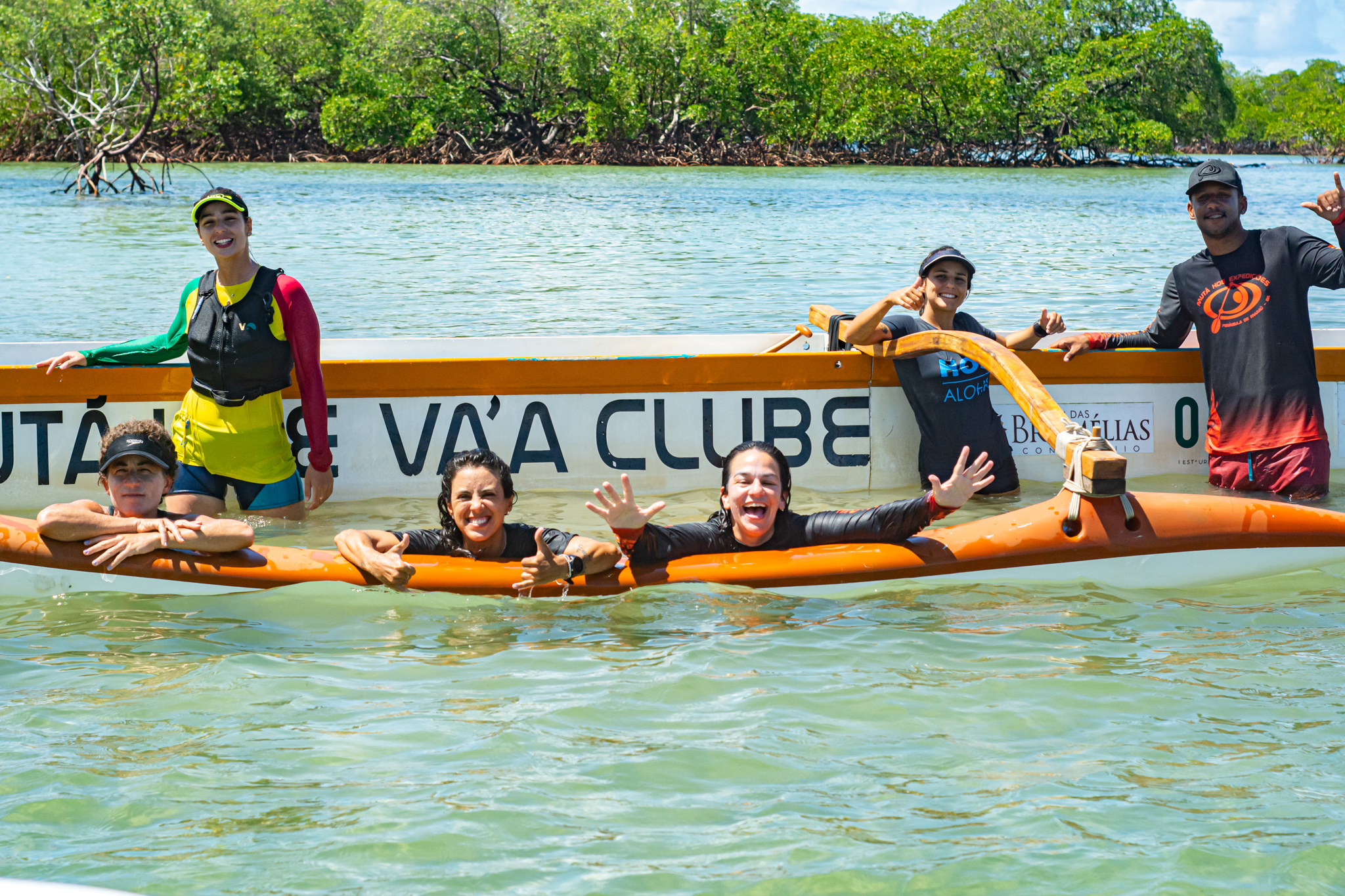 Tripulação sorridente na água ao lado da canoa do clube, com mangue ao fundo